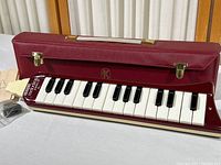 Full view of maroon melodica with piano keys inside open burgundy vinyl case with brass clasps.