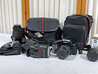 Wide view of all items in the lot showing the camera, lenses, flashes, binoculars, and camera bags arranged on a white surface against a white curtain background.