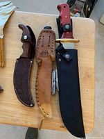 Three knives including a larger fixed-blade knife with red handle in black sheath, smaller wooden handled knife in leather sheath, and folding multitool, on wooden surface.