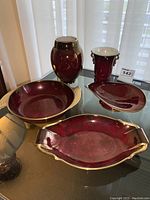 Five Carlton Ware pottery pieces in deep ruby red with gold scroll detail displayed on glass table with curtain background