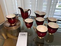 Photo showing 5 Carlton Ware demitasse teacups, 6 matching saucers stacked, one creamer jug, and one sugar bowl in deep ruby red with gold scroll detail.