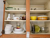 Two-shelf oak kitchen cabinet filled with vintage 1950s kitchenware including casseroles, mixing bowls, glass cups, ceramic seasoning containers and a teapot