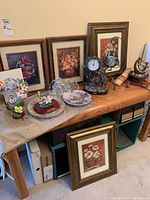Mid-sized oak table with framed vintage decoupage artwork, decorative clock, porcelain flower figurines, and plates arranged on top.