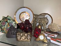 Photo showing the full lot on a glass shelf including hand-painted Chubu dish on the left, brass archway with bell, Buddha figurines (red and dark brown), brass three-monkey sculpture, small clock, metal engraved plates in the back, and textured cream floral carving on the right.