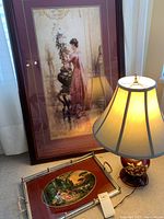 Photo showing framed print, red serving tray with oval scenic inset, and lit pottery base lamp