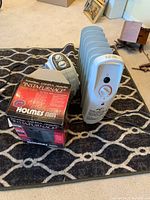 Three heaters on a patterned rug: white oil-filled radiator heater, gray compact heater, and boxed Holmes Insta-Furnace heater