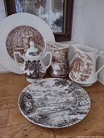 Five pieces of brown transferware displayed on a wooden surface including two large pitchers, one smaller sugar bowl with lid, one large plate, and one smaller plate.