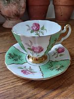 Teacup and saucer set with pink roses and green panels with gold trim on wooden surface.