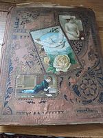 Front cover of the large antique scrapbook with embossed ornate details and pasted vintage illustrations of a boy reading, a woman, a dove, and a rose.