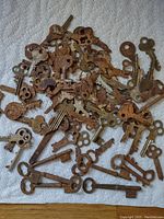 Top down view of a pile of rusty old keys spread on a cloth. Various skeleton and clock keys with diverse sizes and shapes are shown with visible surface rust.
