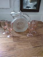 Photo showing clear depression glass jug with circular ridge and floral pattern and four pink depression glass goblets with patterned bowls and stems on wooden surface.