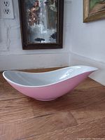 Side view of the pink and white asymmetrical pottery bowl on a wooden surface with a mirror and wall in the background.