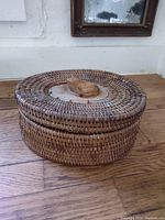 Side profile of round wicker box with lid and carved wooden frog handle on top, placed on wooden surface