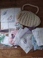 Photo showing the beige knitted ladies purse with a handle, surrounded by assorted embroidered and lace-edged handkerchiefs in different colors and patterns, some in plastic bags.