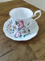 Close-up image of white porcelain teacup and saucer with pink and white Apple Blossom floral pattern and gold trim, placed on wooden surface.
