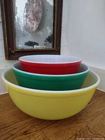Three nested Pyrex mixing bowls in yellow, green, and red on wooden surface, with vintage framed mirror in background.