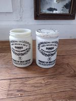 Photo shows two cylindrical white milk glass jars side by side, both labeled with black text and ornate printed design for Dundee Seville Orange Marmalade. One jar has a metal lid with matching print. Jars placed on wooden surface.