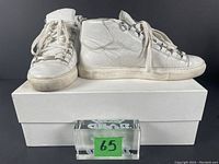 Pair of white leather high top sneakers on white Balenciaga box with green lot number 65 in front. Shoes show signs of wear and dirt on soles and leather.