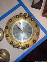 Heavy brass nautical clock with 9 inch diameter designed like a ship’s porthole, showing clock face with dual hour markings and brass round frame with bolts