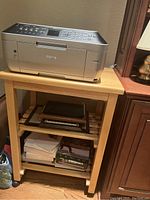 Printer table with a Canon printer on top, showing two slatted shelves with books and framed items, demonstrated rolling wheels, light wood finish.