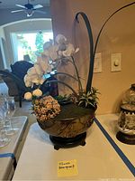 Photo of artificial flower arrangement in copper or brass vase on wooden stand, showing white orchids, brown small flowers, and green leaves.