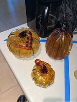 Three blown glass pumpkins with amber color and dark amber stems on a white surface near a coffee maker.