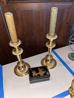 Two brass candlestick holders with beige candles, ribbed bases and top holders on white surface, wooden background.