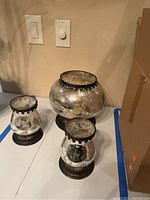 Photo showing three mirrored glass candle holders on table with switches in background, one smaller holder broken