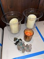 Two glass lantern candle holders each containing a cream-colored artificial candle, Valencia scented candle in a glass decorative holder, and decorative glass grapes on a white surface.
