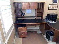 L-shaped desk setup with black desktop surfaces, wooden drawers, shelving hutch, and rolling filing cabinet. Viewed angled in room environment showing usage context.