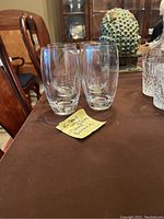 Four clear, heavy glass Tommy Bahama water glasses on a brown table with a yellow note in front describing dimensions and brand.