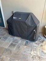 Weber grill covered with black protective branded cover showing full side profile on tiled floor by wall with door and planter close by.