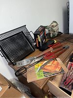 Photo showing an assortment of grill accessories including a cedar grill plank, metal grill brush, wire grill racks, tongs, scissors, a meat thermometer box, serving tools, and a BBQ-themed glove.