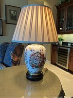 Full view of the porcelain lamp on wooden pedestal with pleated cream lampshade on glass table in living room setting.