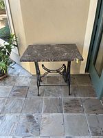 Square dark marble top on an ornate black cast iron Singer sewing machine base, positioned on tiled floor.