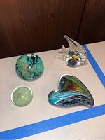 Four blown glass paperweights arranged on a white table with blue tape. Items include heart-shaped and spherical designs with color patterns inside, showing overall lot contents.