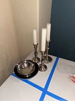 Photo showing three silver toned candle sticks with white candles and one silver serving bowl with black rim on a white surface.