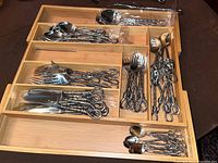Photo showing various silverware pieces arranged neatly in a wooden tray including forks, knives, and spoons with decorative twisted loop handles.
