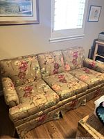 Front angle showing the full floral fabric sofa with three seat cushions and rolled arms, fabric skirt at the base, placed on wooden floor.
