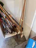 Full view of yard tools leaning against a wall, shows multiple rakes, shovels, and hoes with wooden handles and metal heads.