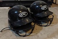 Two black Rawlings baseball helmets with face guards and visible scuff marks, placed side by side on the floor.