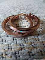 Close-up photo of a pair of large copper hoop earrings showing opal inlay on a textured grey carpet.