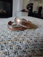 Pair of large copper hoop earrings with etched Greek key pattern and rhinestones on one edge, laid on a light colored textured fabric surface.