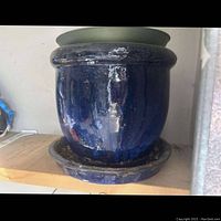 Side view of large deep blue glazed ceramic planter sitting on a wooden shelf with matching round bottom tray beneath it. Shows glossy reflective finish and light grime.