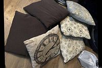 Six decorative pillows arranged on the floor showing variety of patterns and colors: two solid brown, one clock design, one floral, two circular pattern.