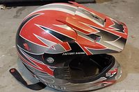 Side angle view showing red, black, and silver design with 'Factory Racing' and 'RAIDER' logos. Helmet interior and chin strap visible.