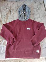 Front view of red Roots hoodie with salt and pepper patterned hood laid flat showing front pocket and small Roots patch label.