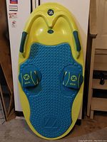 Full view of yellow and blue water ski training board leaning against a wall, showing handles, foot straps with ZUP branding, and textured foot pad.