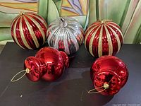 Five vintage Christmas ornaments, three large round striped ornaments and two smaller red bulb-shaped ornaments arranged on a dark surface against a painted backdrop.