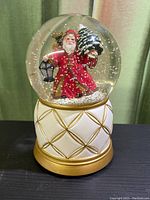 Snow globe showing Santa Claus inside glass globe holding Christmas tree and lantern on raised white and gold base